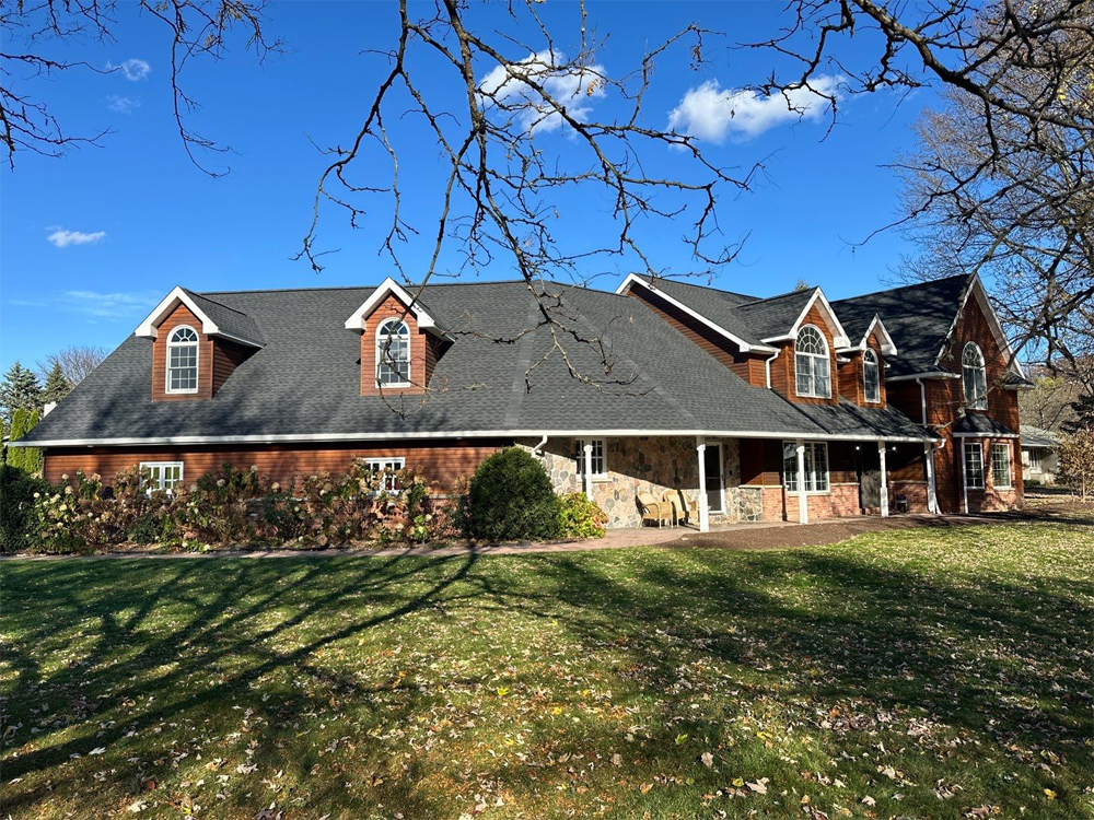 Large brick and stone home with a new dark asphalt shingle roof under a clear blue sky in a residential neighborhood.
