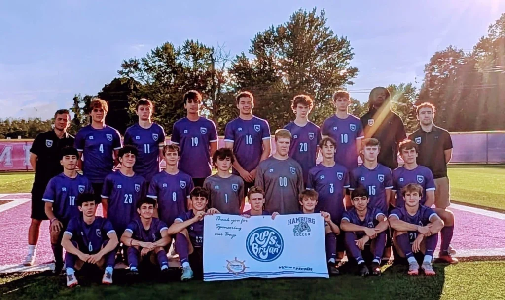 Hamburg soccer team posing with a Roofs by Bryan sponsorship banner on a field during a community event