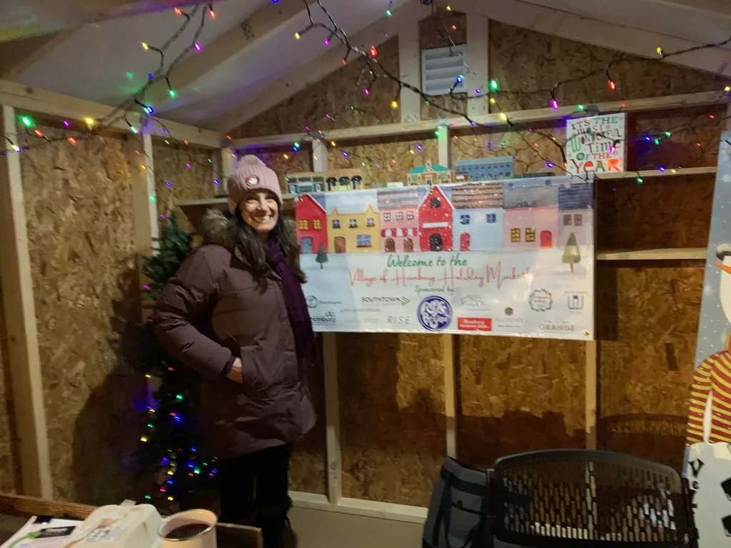 Woman in winter coat and hat standing inside a decorated holiday market booth with colorful string lights and festive sign