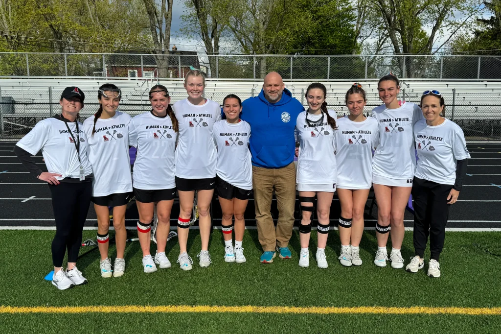 Girls lacrosse team and coaches wearing "Human Athlete" shirts posing together on a field in front of bleachers