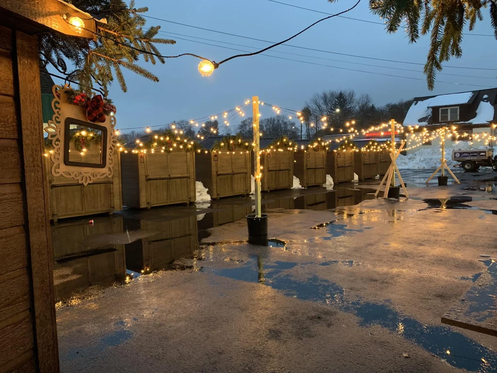 Outdoor holiday market stalls decorated with string lights at dusk with wet pavement and snow in the background