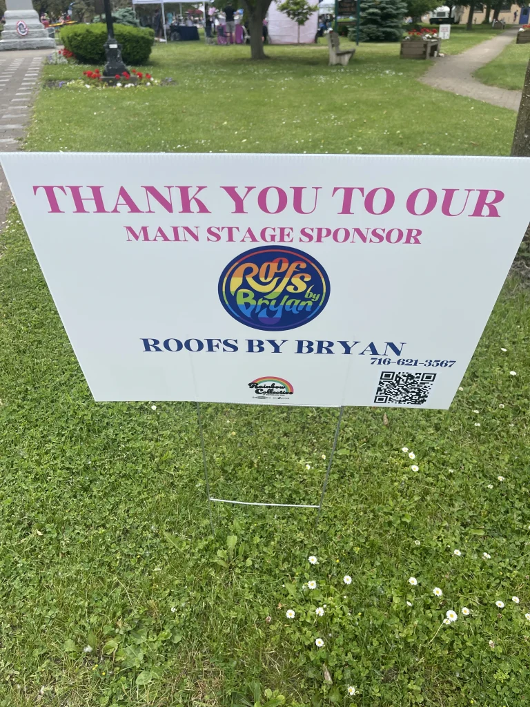 Roofs by Bryan main stage sponsor sign displayed at a community event in a park with tents and greenery in the background