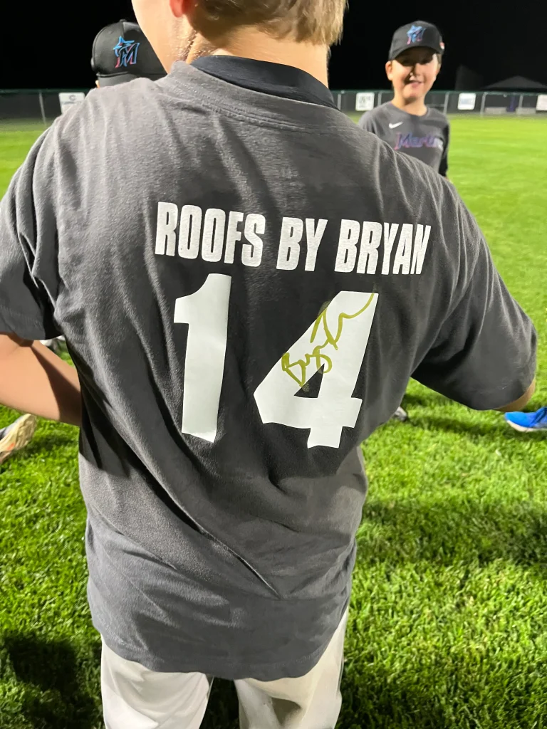 Child wearing a "Roofs by Bryan" baseball jersey with number 14 on a field at night during a youth baseball practice