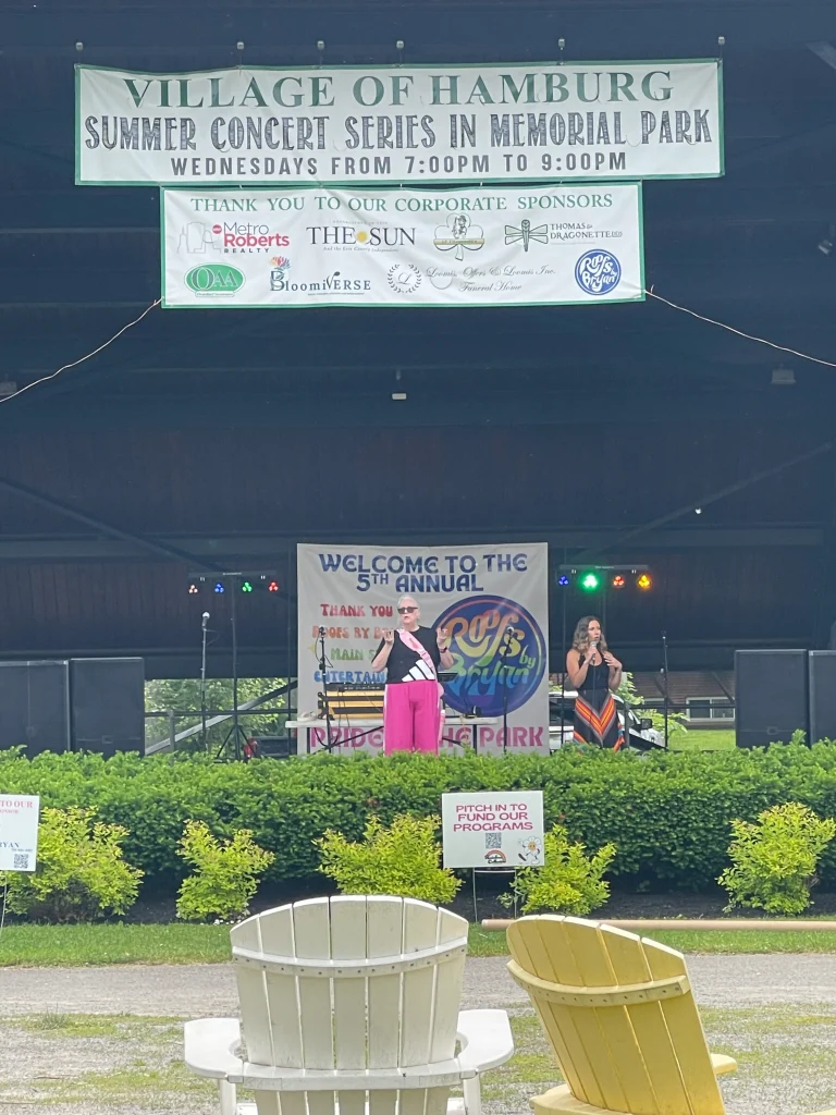 Stage at Village of Hamburg Summer Concert Series in Memorial Park with performers and signage, sponsored by Roofs by Bryan
